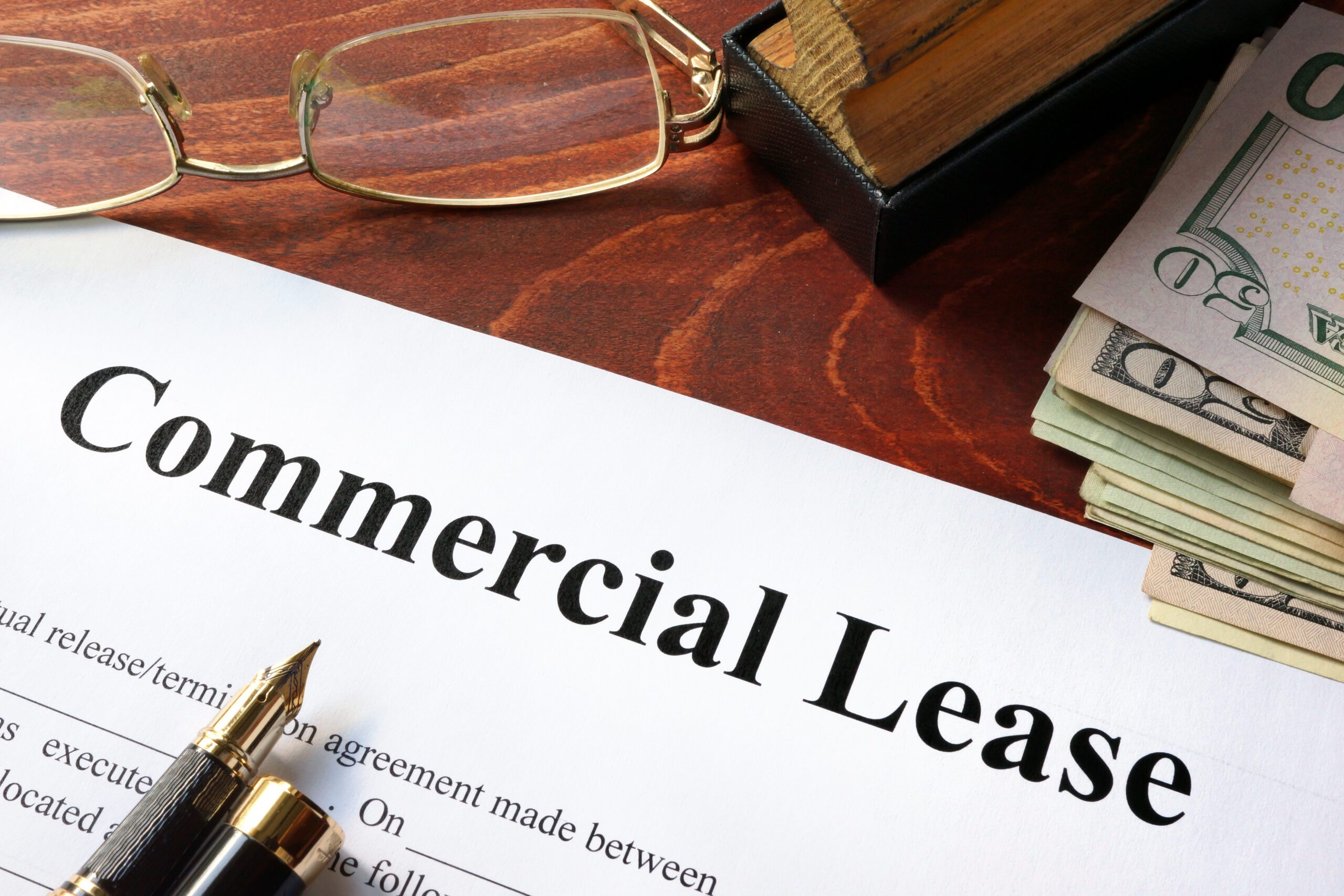An image of a piece of paper that reads “commercial lease” with a pen, glasses, a stamp, and a stack of 50-dollar bills in USD.