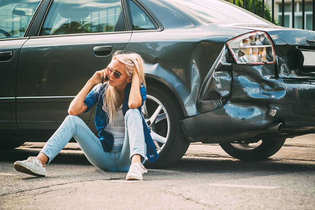 A blonde woman sits on the ground, on the phone, next to her car with its rear end smashed in.