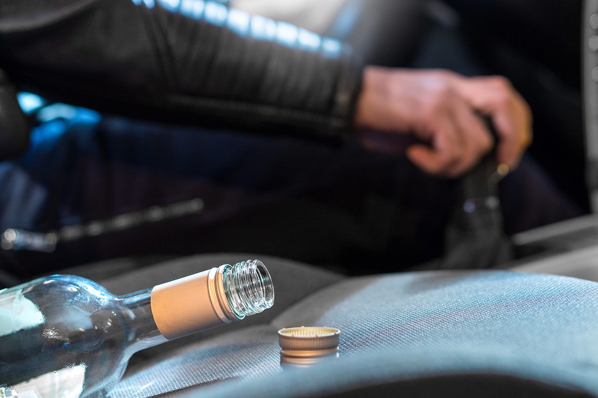 An empty, open wine bottle is open in the foreground while the driver has their hand on the shifter.