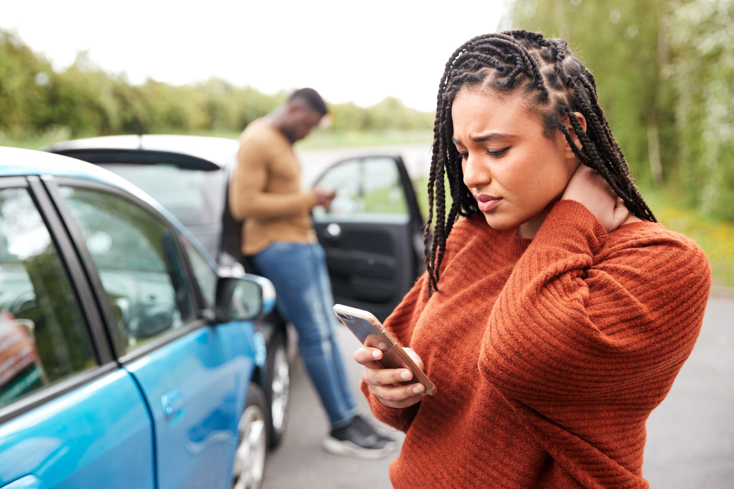 Two people, a man in the background and a woman with her hand on the back of her neck in the foreground, are standing in front of a car accident, looking at their phone screens.