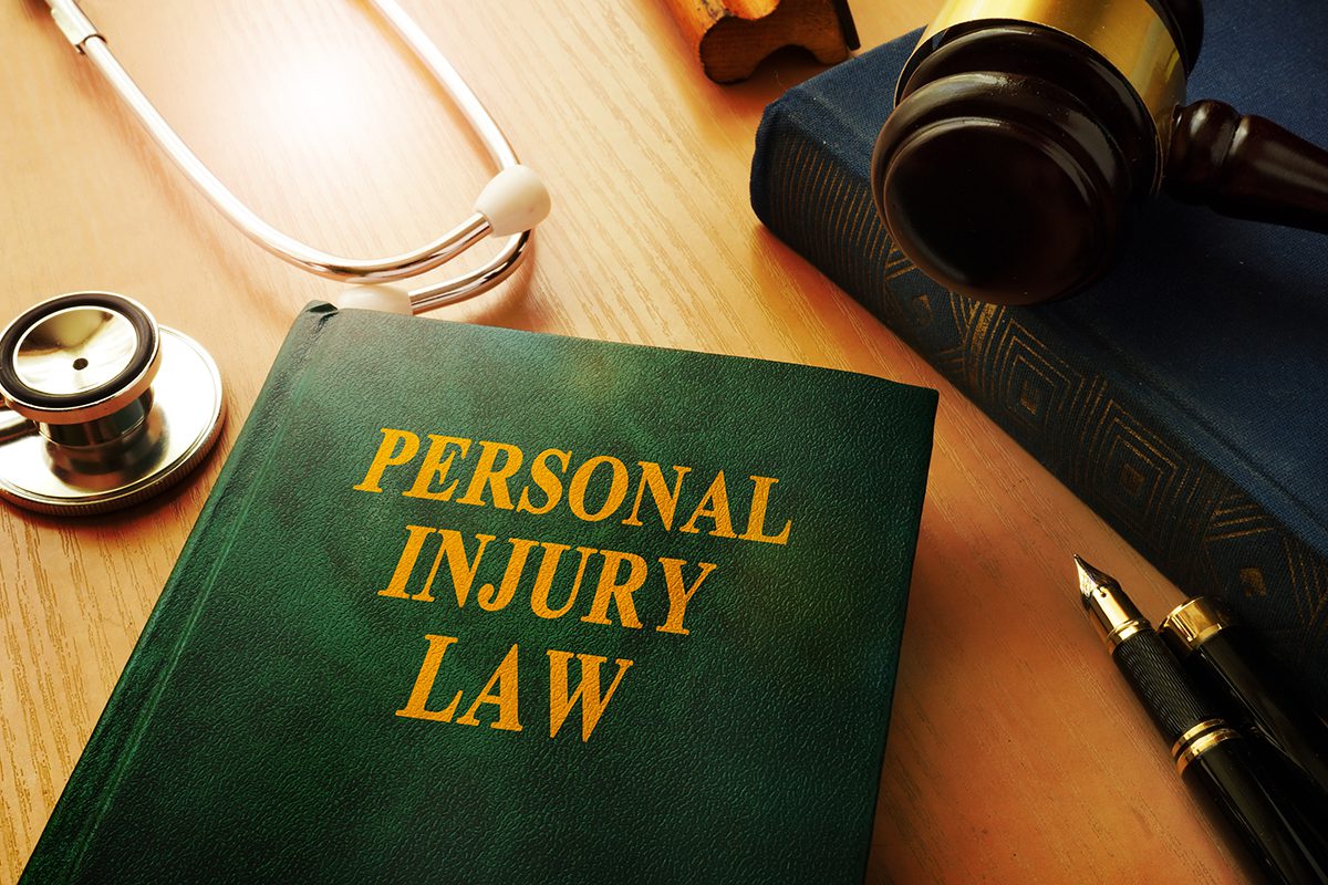 A green book with yellow writing on it that says “personal injury law” is surrounded by a stethoscope, a pen, and a blue book with a gavel resting on it.